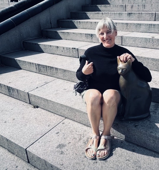 Lincoln based artist Zoe Haith sits with her dog, on some large stone steps. The image leans heavily into dark and light mono colours with Zoe wearing a black top and shorts, with the black shadows on the steps filtering across the screen, against a light grey contrast.